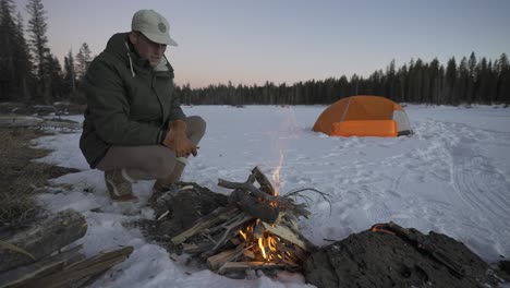 man feeding a fire in the snow next to his tent