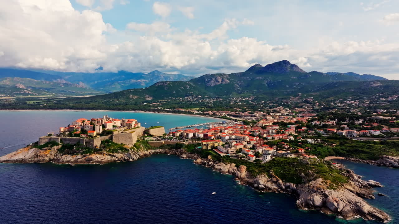 Aerial drone shot over the historic old town of Calvi, Corsica, France. Picturesque view of coastal town, landscape, fluffy clouds, and mountains in the background