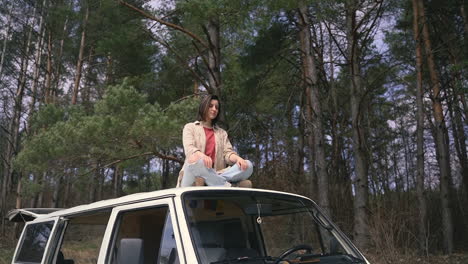 una linda morena está sentada relajada sobre el techo de una caravana en medio del bosque