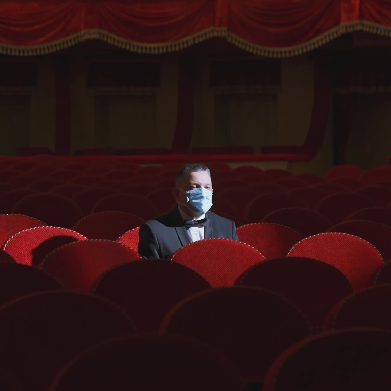 Rich man among rows of empty red chairs. Businessman in medical mask sitting in comfortable chairs alone on dark theater background. Pandemic in the world.
