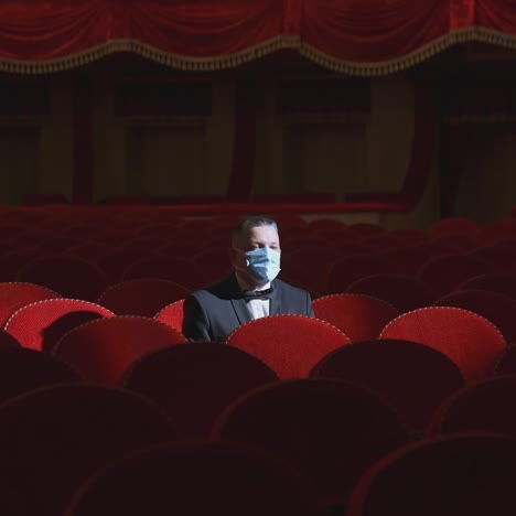 rich man among rows of empty red chairs. businessman in medical mask sitting in comfortable chairs alone on dark theater background. pandemic in the world.