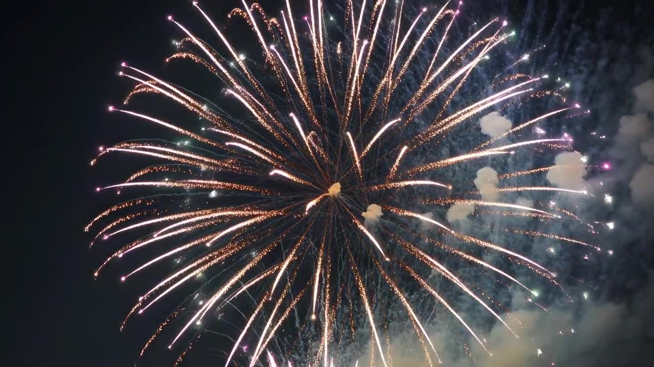 Shell bursting aerial firework, sending white and gold trails at public display, with smoke plume