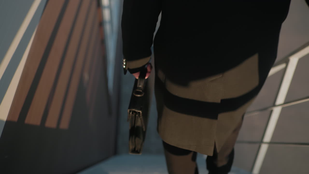 뒷면으로 가까이 보기: 검은 코트 남자가 계단을 내려가고, 서류 가방을 들고, 배경의 건축물, 천에 반사되는 줄무, 계단 케이스의 철 레일링을 들고 있습니다.