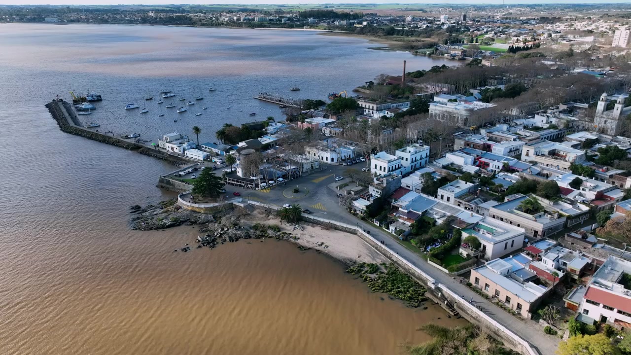 Avanzamento sul lungomare di Colonia del Sacramento che rivela edifici coloniali imbiancati, l'area del porto e barche ormeggiate lungo la costa uruguaiana
