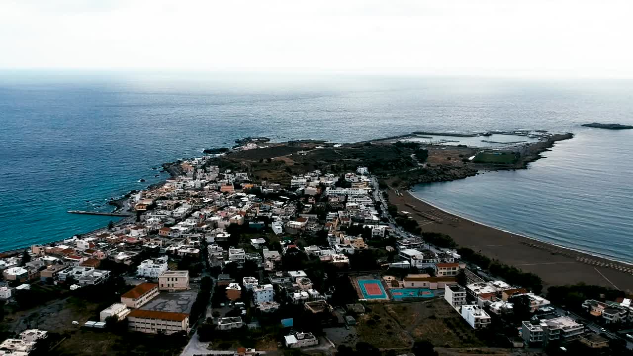 Aerial drone view of quiet coastal town built on narrow peninsula with calm sea on both sides. Beach, houses, and sports courts visible under cloudy sky in peaceful seaside setting.