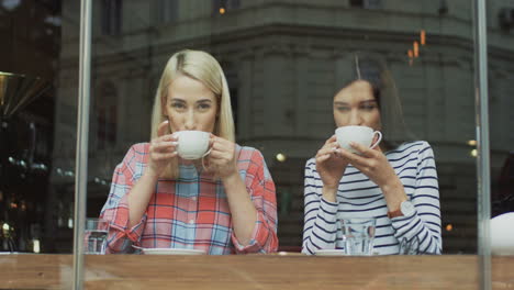 Portrait-Shot-Of-A-Blonde-And-Brunette-Girls-Spending-Their-Morning-Together-And-Having-A-Conversation-While-Drinking-Coffee-In-A-Cafe