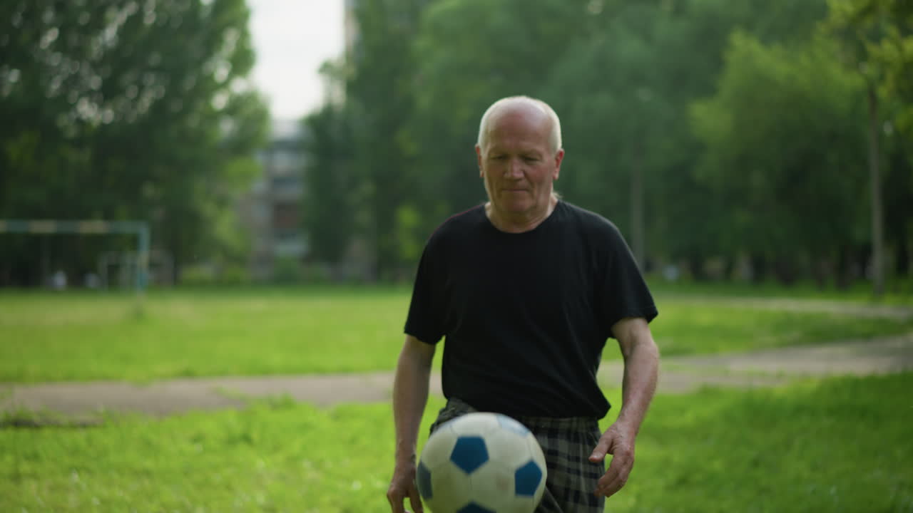 An elderly man skillfully taps a soccer ball up in a lush grassy field, surrounded by trees and a goalpost in the background, focused as he taps the ball and kicks it