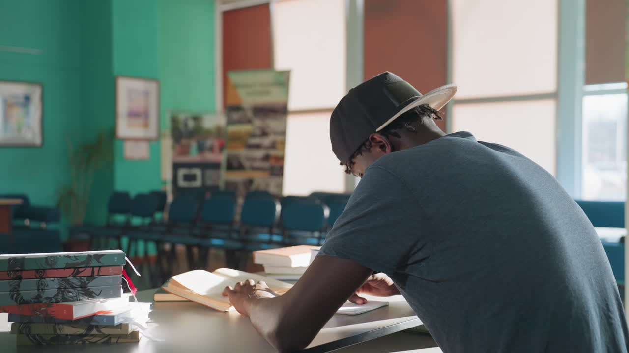 Rear view of student sitting at desk in sunlit library, writing notes in open book with pen, surrounded by stacked books, focused on study in quiet room decorated with framed artwork
