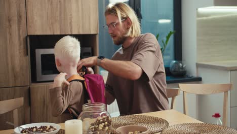 Ein-Blonder-Mann-Mit-Bart-Und-Brille-Legt-Seinem-Kleinen-Albino-Sohn-Mit-Weißen-Haaren-Einen-Leuchtend-Roten-Rucksack-An-Und-Sagt-Ihm-Nette-Worte,-Bevor-Ein-Neuer-Schultag-In-Der-Schule-Beginnt