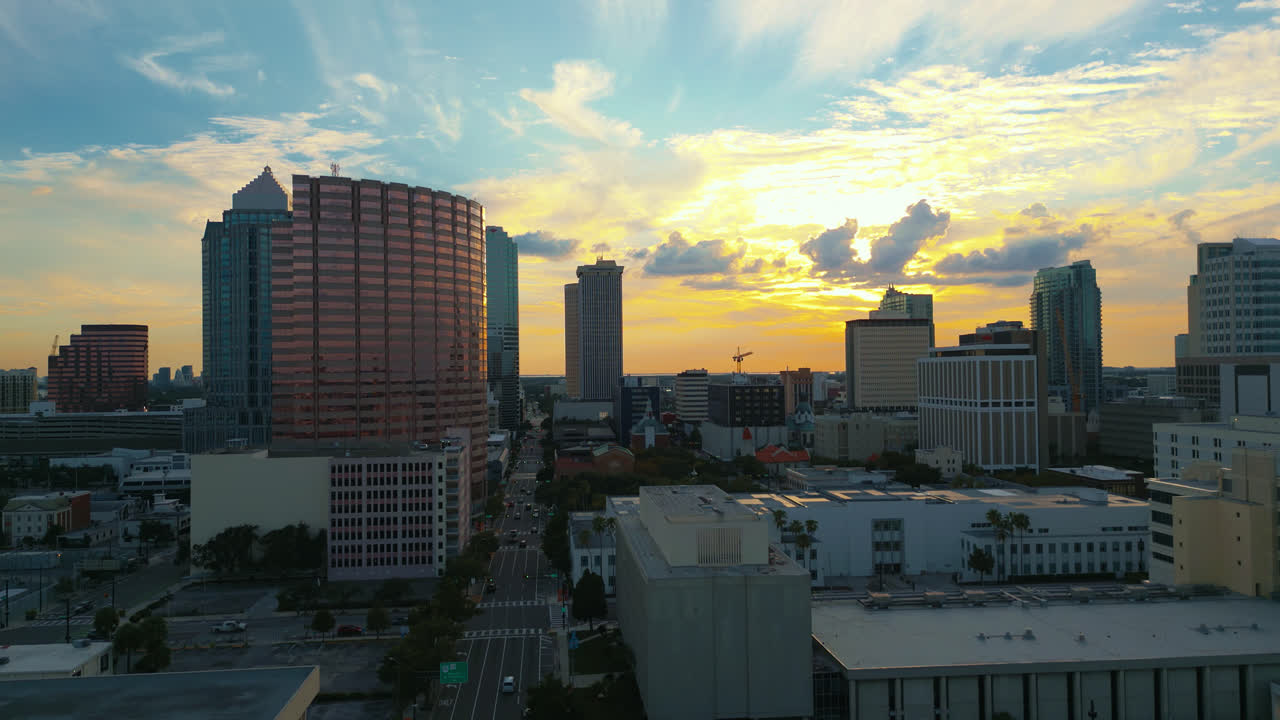 Aerial rising shot of the Tampa skyline with the sun setting behind the clouds as blues and yellow fill the sky and busy cars drive along the streets