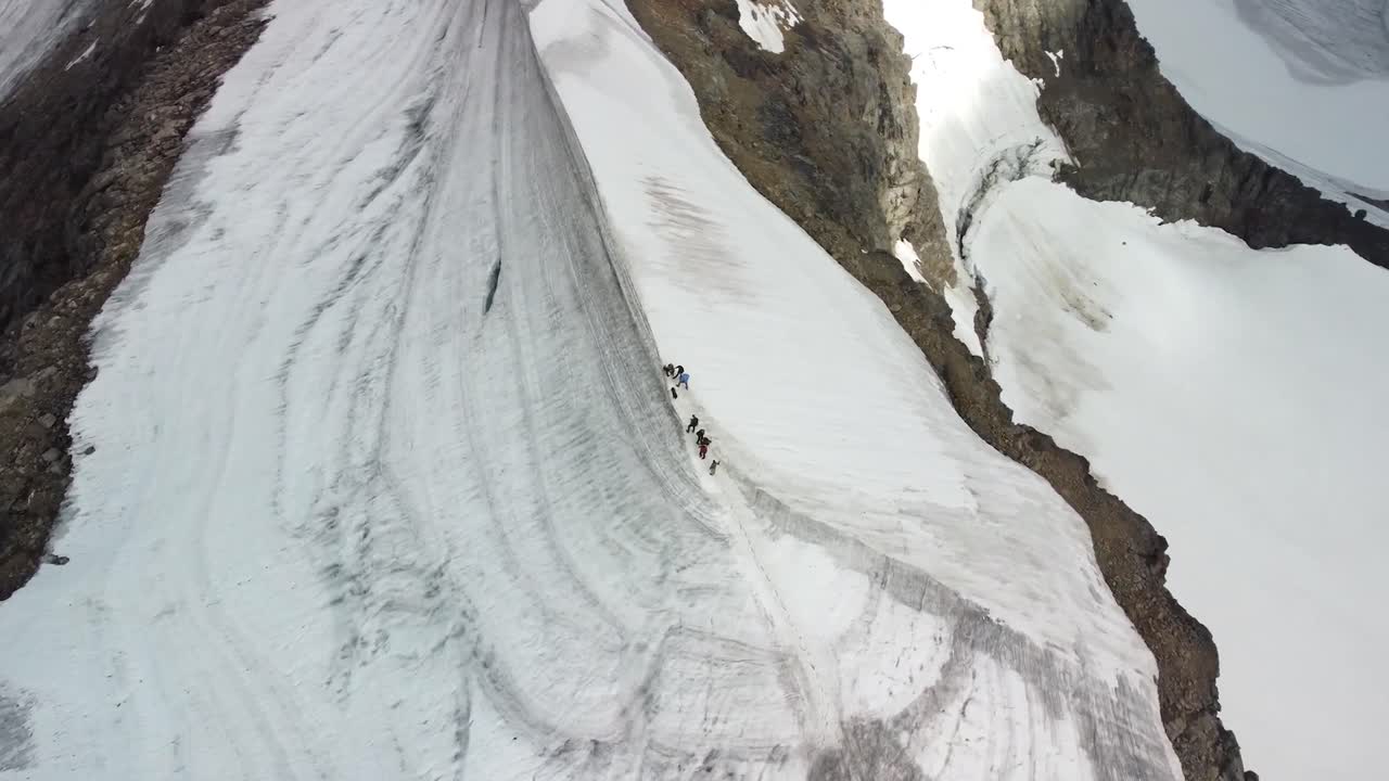 Top down aerial drone footage orbiting and spinning around a group of people who are walking and standing on top of a white snow covered mountain edge during a sunny day. Rocky cliffs visible on sides