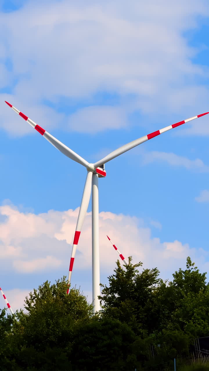 Wind turbine with red lines on the blades rotates in the wind. Green trees grow under the windmill. Low angle view. Vertical video.
