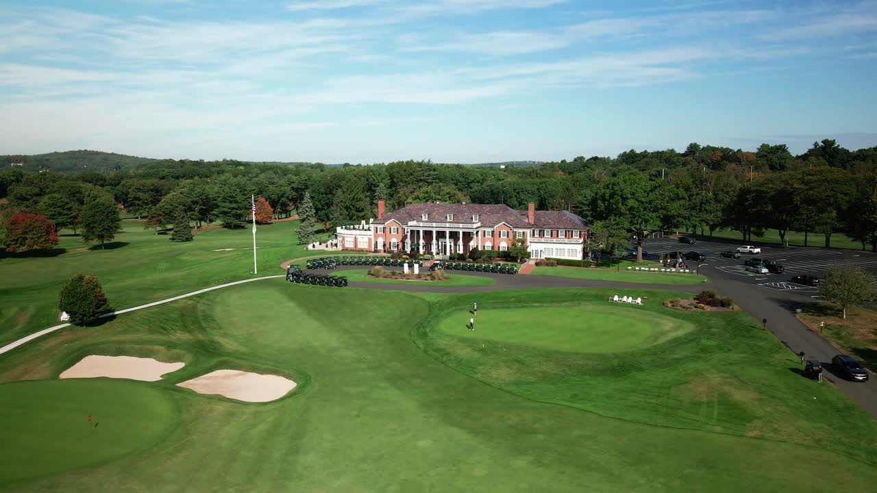 Dronebeelden tonen een elegant clubhuis in koloniale baksteenstijl op een prestigieuze golfbaan met verzorgde greens, zandbunkers en opgestelde golfkarretjes.
