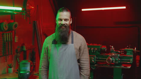 portrait of caucasian young male welder with long beard and wearing apron turning face to camera and smiling cheerfully in workshop