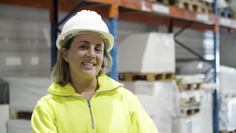 mujer rubia caucásica con casco y uniforme parada en el almacén y sonriendo a la cámara