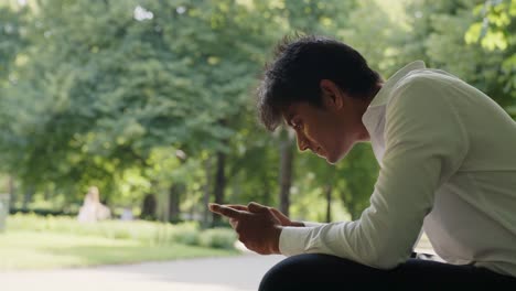 Lächelnder-Asiatischer-Kerl,-Der-An-Einem-Sonnigen-Tag-Mit-Dem-Handy-Im-Park-Plaudert