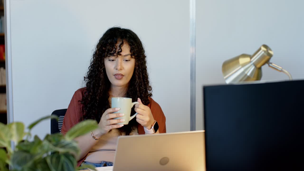 African American woman in rust cardigan staring at laptop, sipping mug, reading screen, working