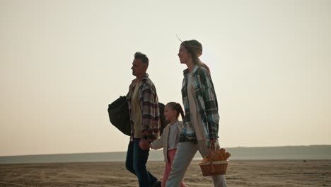 Side-view-of-a-happy-brunette-girl-in-a-green-checkered-shirt-walking-and-holding-hands-with-her-little-daughter-and-husband-during-her-hike-and-picnic-in-the-summer