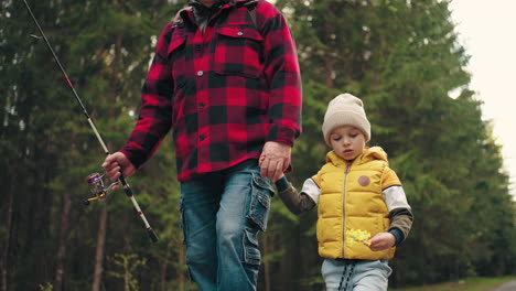 cute little boy and his granddad are walking together in nature strolling on road across forest going to fishing