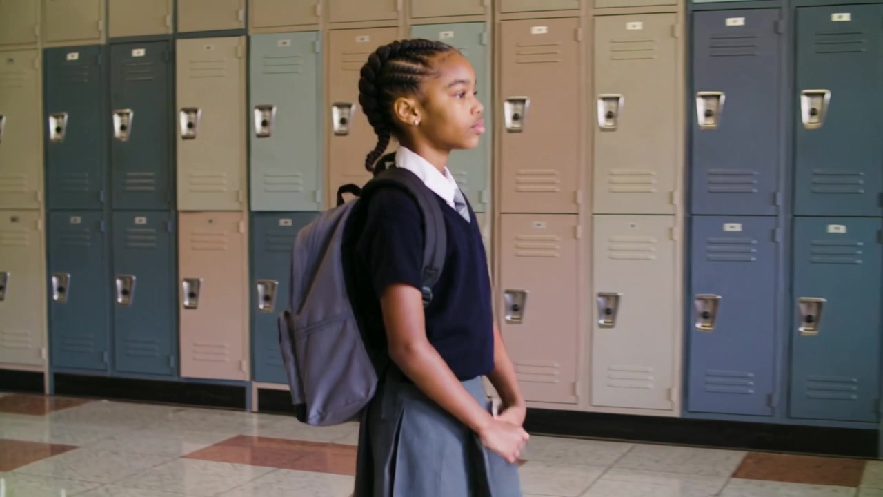 Shifting weight, student in uniform walking along metal locker bank at school with gray backpack