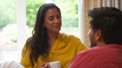 couple relaxing sitting on sofa at home together with hot drinks talking