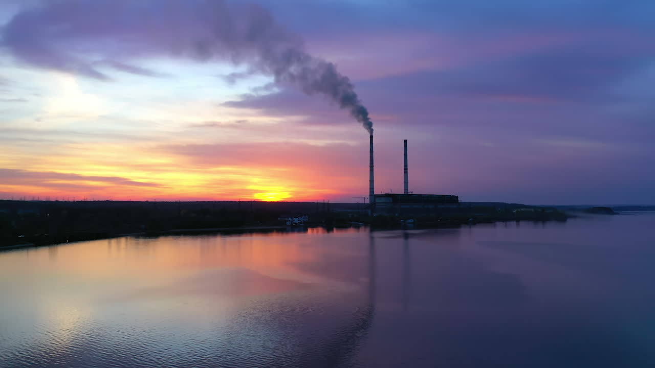 Chemical plant over the river at sunset. Harmful smoke released in the air from industrial factory in the evening. Camera rising up.