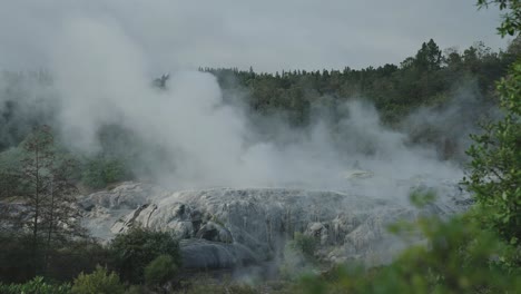 Geothermal-geyser-Erupting-with-steam-and-water-surrounded-by-nature,-Rotorua,-New-Zealand,-Slow-motion-iconic-steamy-rocky-environment,-Sunny-Daytime-Sky