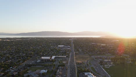 Overhead-flight-above-downtown-city-expressway-and-skyline-towards-water-and-mountain-range-with-bright-white-sun-in-background,-aerial-approach