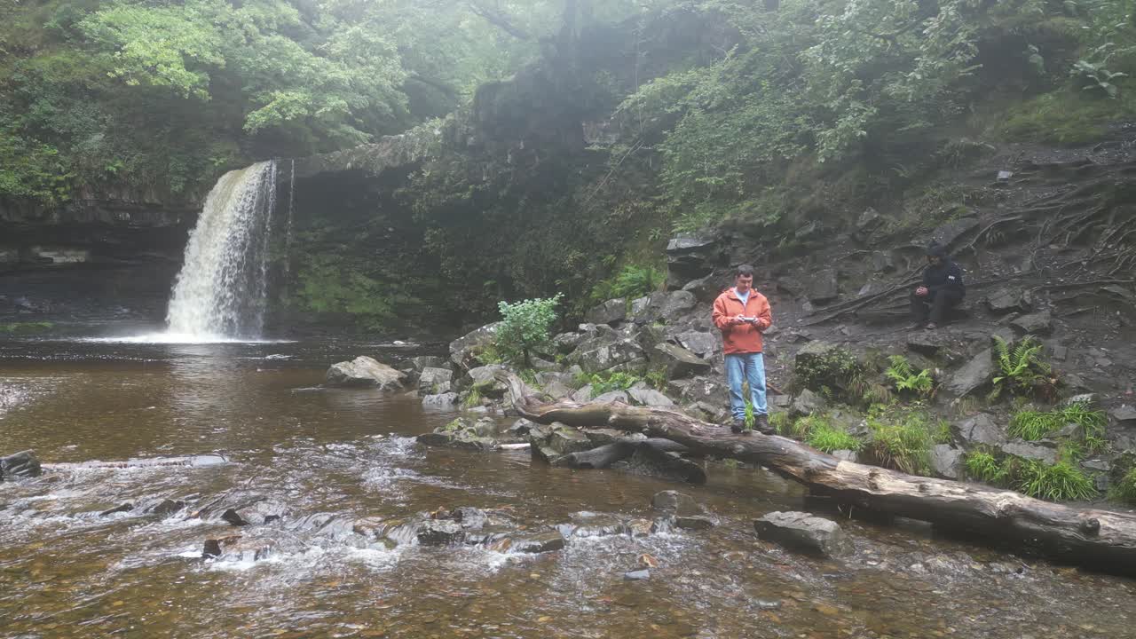 Drone footage of a young man standing by Angel Falls in Brecon Beacons National Park on a cold, wet day, gazing away from the cascading waterfall, surrounded by rugged rocks and lush green forest