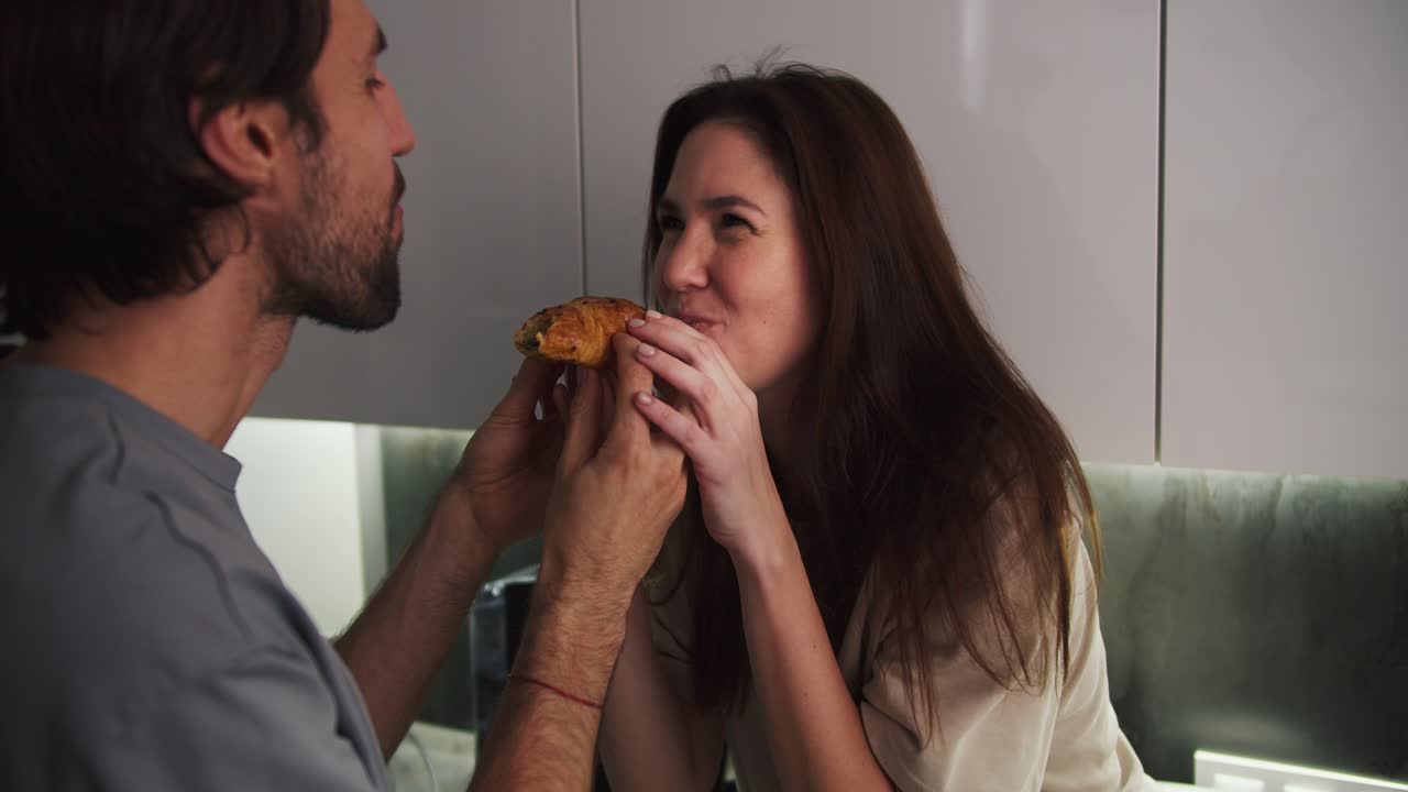 un feliz chico moreno con paja junto con su novia morena en una camiseta beige simultáneamente morder un croissant de diferentes lados durante una cita romántica en la cocina por la noche