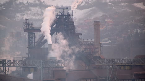 Smoke-rises-from-industrial-structures-amidst-a-snowy-mountain-backdrop,-highlighting-the-stark-contrast-between-industry-and-nature