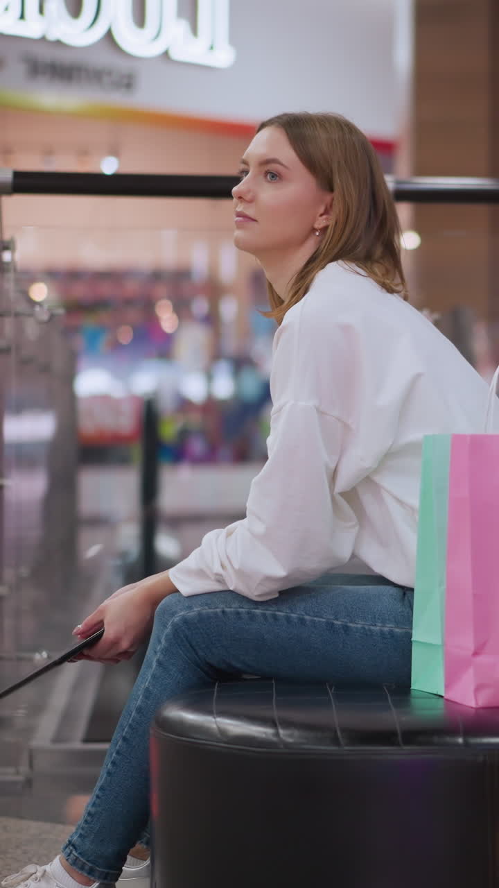 mujer joven sentada en un banco en un centro comercial sosteniendo una tableta, rodeada de iluminación y decoración vibrantes, bolsas de compras brillantes a su lado y atmósfera festiva