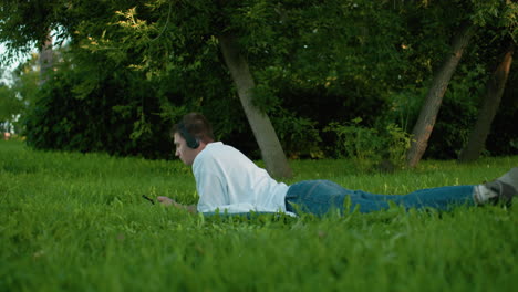 man lying on grassy field wearing headphones, white top, and jeans, nodding his head to music, surrounded by lush greenery and trees, enjoying a serene outdoor moment