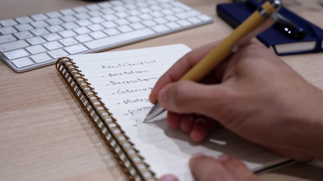 Close-up of a hand writing an event checklist in a spiral notebook on a wooden office desk with keyboard, captured in soft indoor light with shallow depth of field