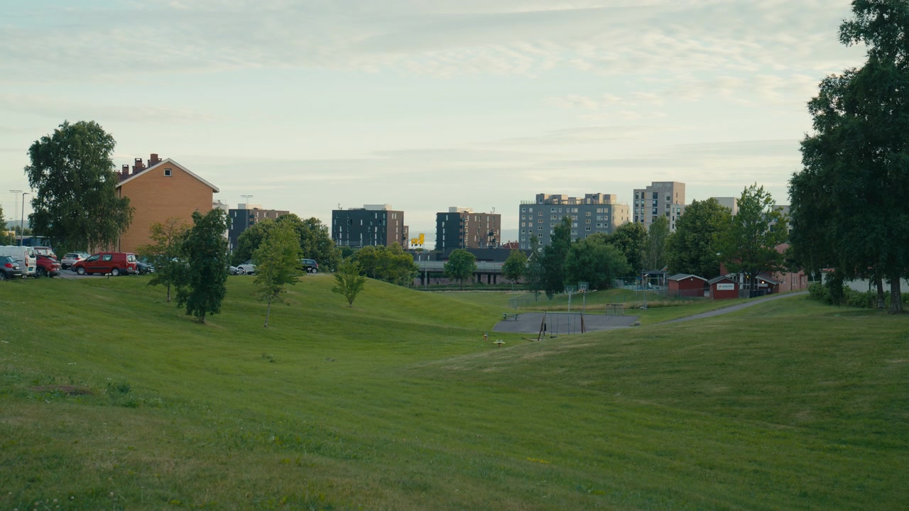 Palazzi residenziali si ergono oltre l'erba ondulata, gli alberi sparsi e un tranquillo parco di quartiere a Oslo, Norvegia, creando uno skyline urbano delicato con un'atmosfera suburbana distintamente scandinava