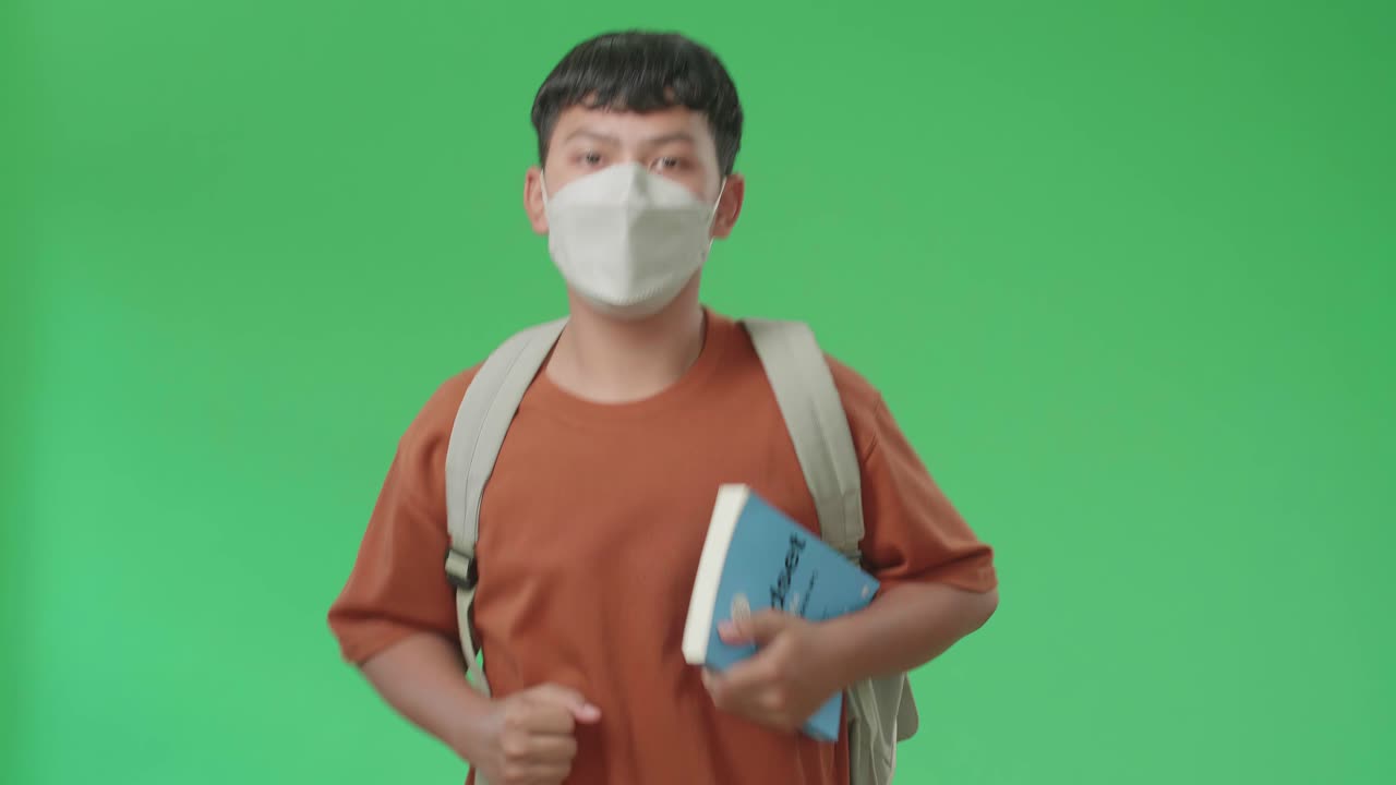 The Front View Of Asian Boy Student Wearing A Mask, Backpack And Brown T-Shirt Holding A Book And Running To School On Green Screen Chroma Key.