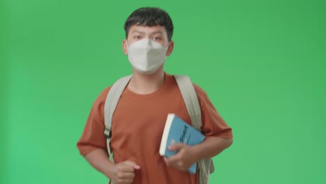 the front view of asian boy student wearing a mask, backpack and brown t-shirt holding a book and running to school on green screen chroma key.