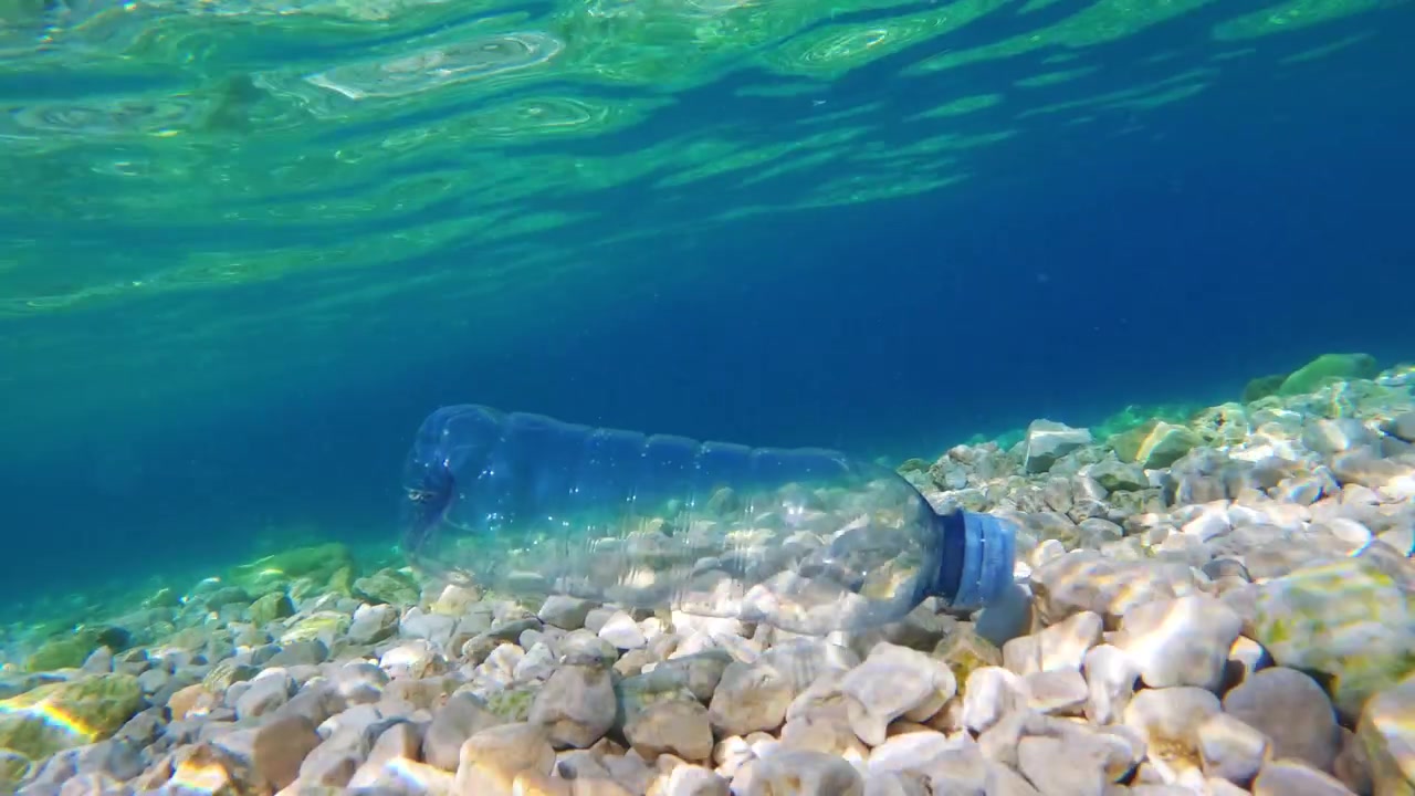 Uma garrafa de plástico repousa no fundo do oceano entre pequenas rochas. A água flui acima da garrafa. Este momento capta o contínuo lixo plástico marinho no oceano.