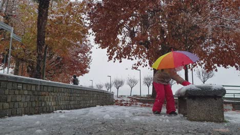 child girl with colored umbrella and red trousers plays kicking snow