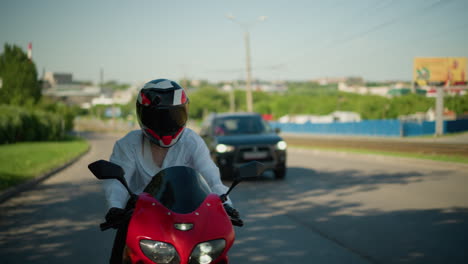 una motociclista femenina en una bicicleta eléctrica roja viaja a través de un área urbana, su camisa abierta por el viento, revelando su singlet y cuerpo, se ve un coche siguiendo por detrás