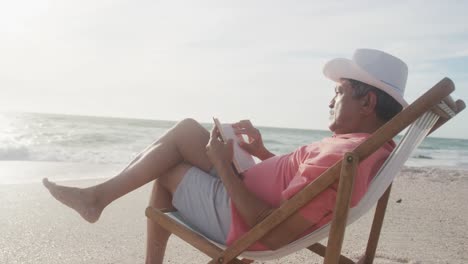 hombre latino senior relajándose en una cama de sol en la playa al atardecer, leyendo un libro