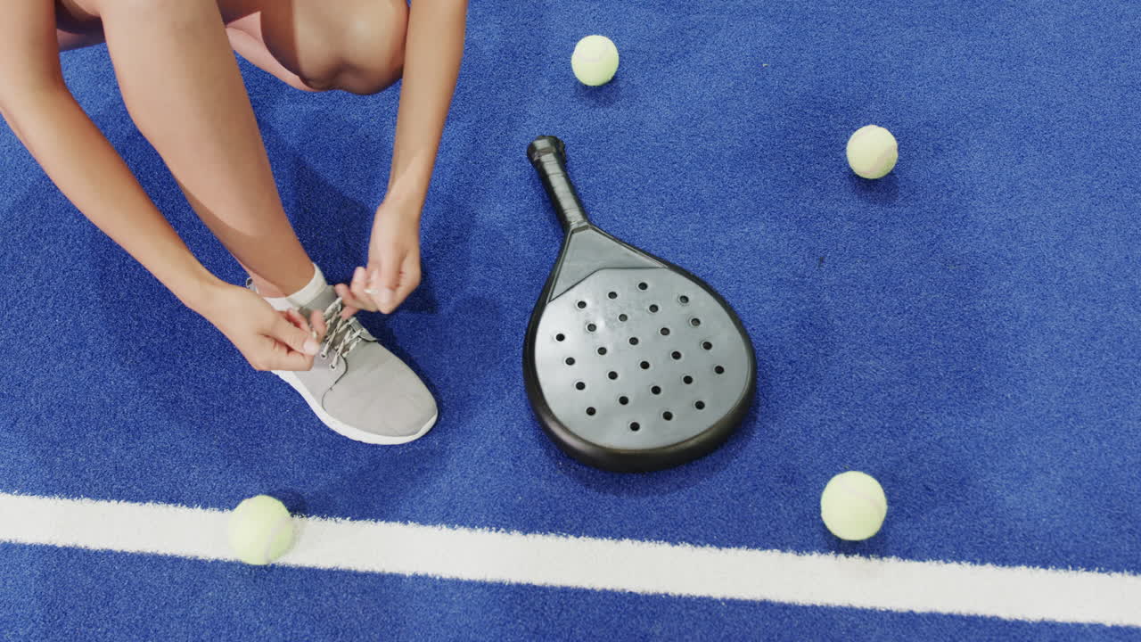 Tying shoelaces on blue court with padel racket and tennis balls scattered