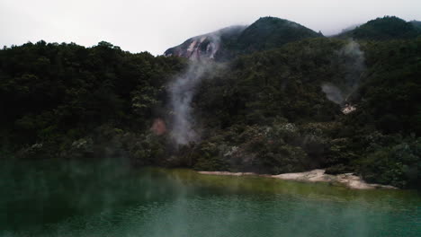 Fly-through-steam-rising-over-geothermal-lake-and-lush-forest-in-New-Zealand,-North-Island