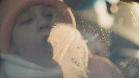 a close-up of a young girl wearing a pink knit hat and white scarf, blowing air onto a foggy car window. her breath creates condensation on the glass