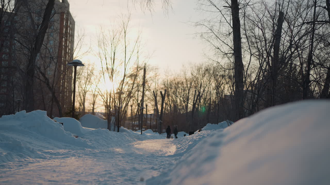 Winter season with two people walking down snowy path lined with street lamps and bare trees, tall buildings in background, soft sunlight, calm winter city park atmosphere with shadows