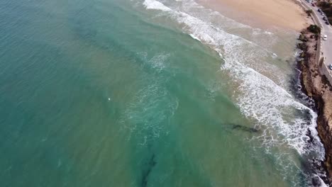 Las-Olas-Verdes-Se-Adentran-En-Una-Hermosa-Bahía-De-Arena-Con-Un-Pueblo-Pintoresco,-Clima-Soleado