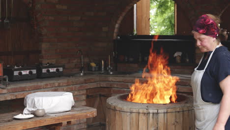 una panadera georgiana parada al lado del horno de arcilla de piedra redonda con fuego ardiente