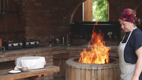 a georgian woman baker standing next to the round stone clay oven with burning fire