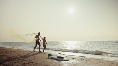 A-happy-couple,-a-blond-guy-in-shorts-and-his-blonde-girlfriend-in-an-orange-swimsuit-are-running-along-the-seashore-and-holding-yellow-and-green-fireworks-in-their-hands-that-leave-a-trail-of-smoke-in-the-morning-on-the-seashore