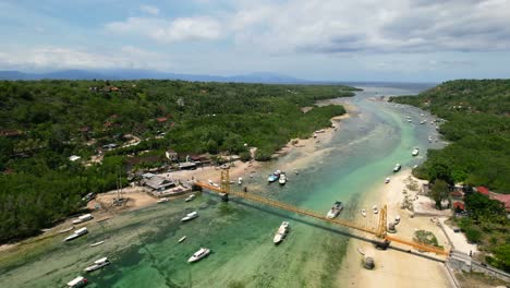 Zoom-Aéreo-Fuera-Del-Puente-Amarillo-Sobre-El-Canal-Entre-Nusa-Ceningan-Y-La-Isla-Lembongan-En-Marea-Baja-Con-Barcos-Anclados-En-Aguas-Turquesas-Tropicales-En-Bali